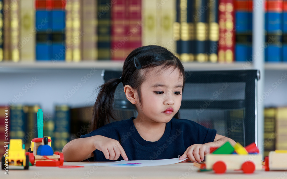 Small pretty pigtails hair preschooler kindergarten happy girl sit on chair drawing with colored pencils on paper on table full of plastic truck toys in living room at home in front bookshelf Small pretty pigtails hair preschooler kindergarten happy girl sit on chair drawing with colored pencils on paper on table full of plastic truck toys in living room at home in front bookshelf