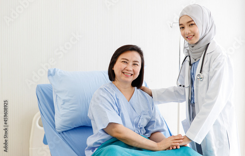 Muslim Arab Islam female doctor wears grey hijab and white lab coat uniform with stethoscope standing look at camera visiting encourage cheering up senior asian woman patient on hospital bed