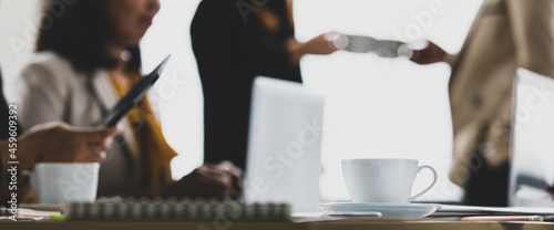 Close up shot of white hot coffee cup and dish on working meeting desk while unidentified unrecognizable businesswoman in casual business clothing working and cheers celebrating in blurred background