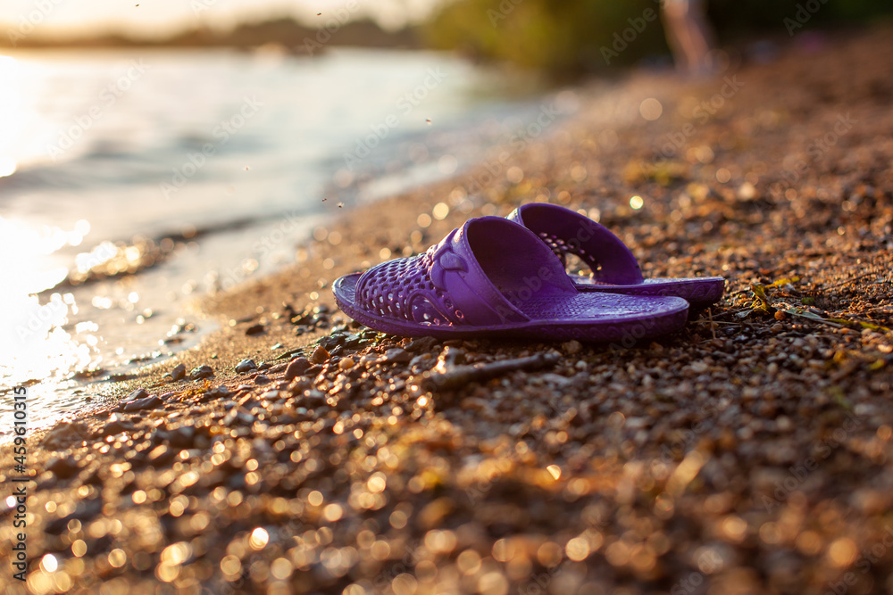 Close-up of rubber slippers on the shore of a lake or river made of small stones against a colorful sunset background. A beautiful place in nature for family holidays and swimming. Close-up of rubber slippers on the shore of a lake or river made of small stones against a colorful sunset background. A beautiful place in nature for family holidays and swimming.