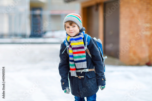 Little school kid boy of elementary class walking to school during snowfall. Happy child having fun and playing with first snow. Student with backpack or satchel in colorful winter clothes.