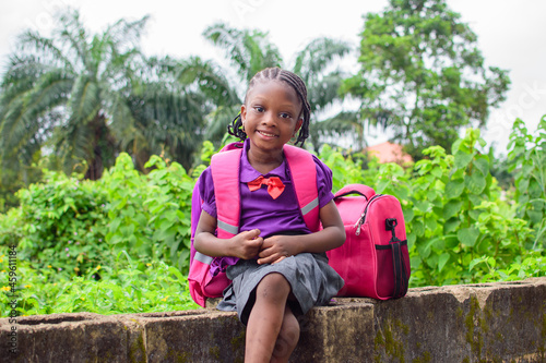 An African girl child in school uniform and with a backpack, sitting outside on a low fence