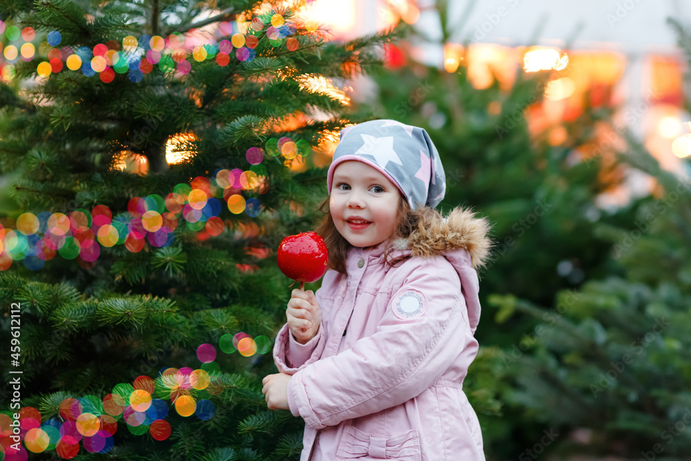 Cute little smiling preschool girl on German Christmas market. Happy child in winter clothes eating sweet sugared glazed xmas apple on with lights on background. Family, tradition, celebration concept