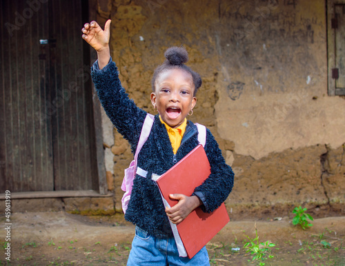 A happy little African girl child or student with pink school bag, holding and hugging her books outside a village mud house