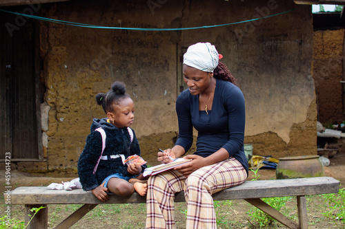 An African mother or teacher outside a village house, helping a girl child with her studies for excellence in her education