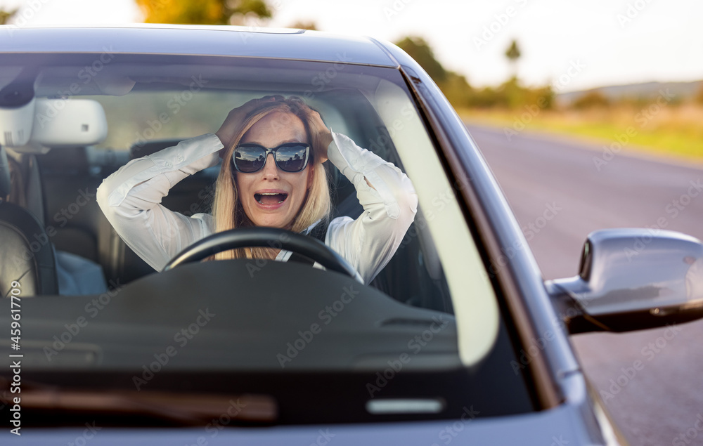Shocked caucasian woman in sunglasses sitting in car with open mouth. Female driver grabbing her head and screaming during accident on road.