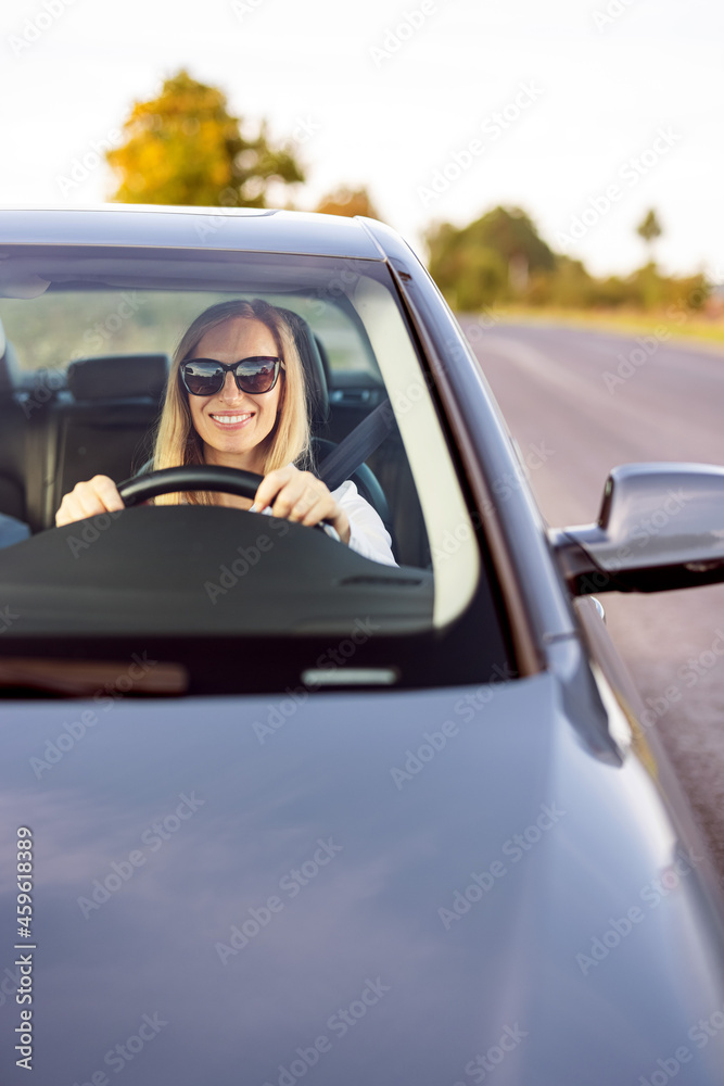 Happy mature woman in formal blue shirt and sunglasses, fastening with safety belt, driving modern black car. Experienced and confident female driver.