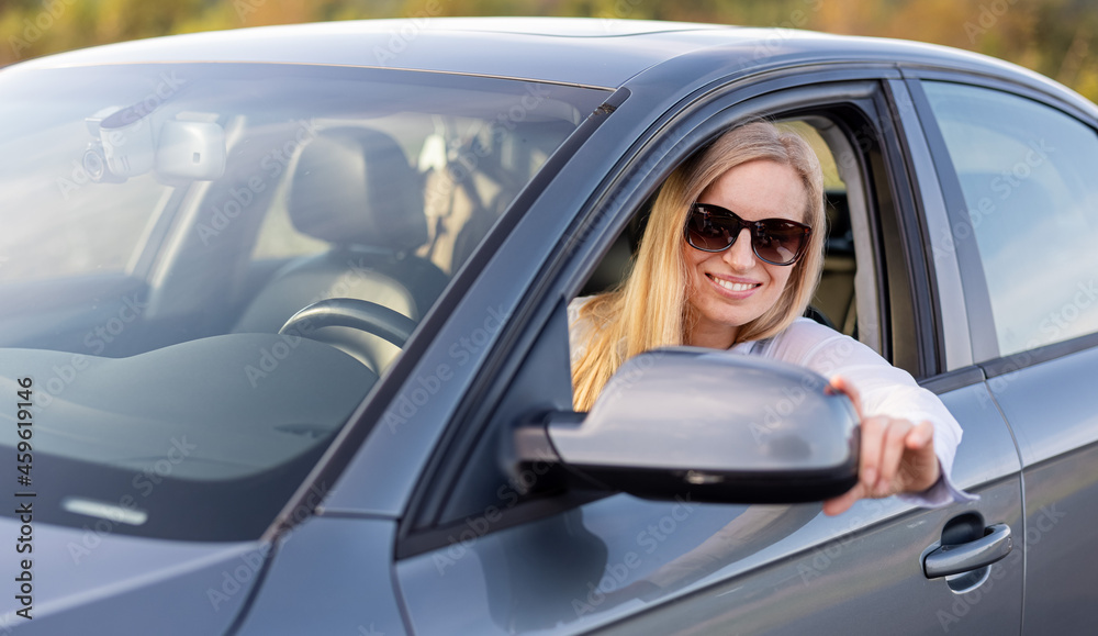 Positive mature lady with blond hair in sunglasses and blue shirt adjusting side mirror before driving car. Concept of people and transport.