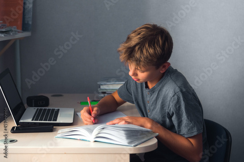 student teenager boy doing homework with laptop, open copybook  and computer, workplace at home photo