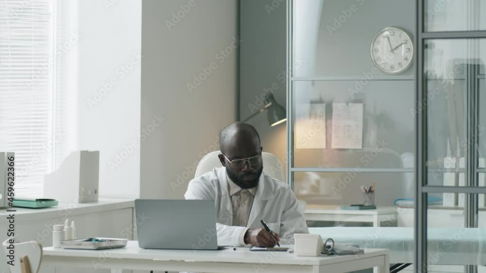 African American male doctor in lab coat and glasses working on laptop and taking notes at desk in medical office African American male doctor in lab coat and glasses working on laptop and taking notes at desk in medical office