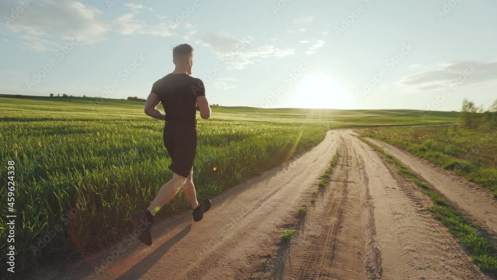 A sporty man is running along a dirt road near a field. He's wearing a sports uniform. Sunset. 4K A sporty man is running along a dirt road near a field. He's wearing a sports uniform. Sunset. 4K