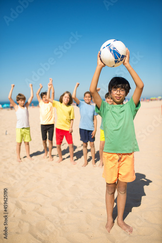 Happy Asian preteen boy posing with ball at beach. Group of excited multiethnic friends playing football at seaside. Summer vacation, friendship concept