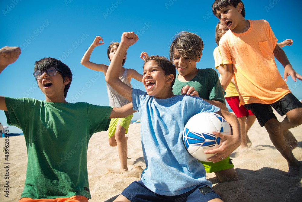 Obraz premium Excited preteen boys cheering after game on beach. Multiethnic friends playing football at seaside. Summer vacation and friendship concept