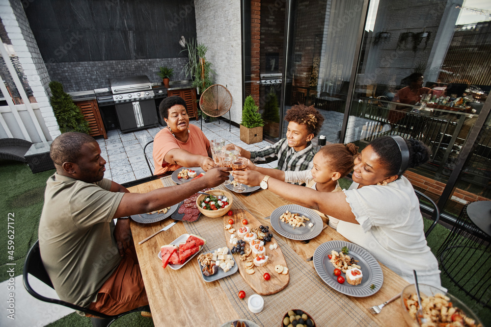 Portrait of big African-American family clinking glasses while enjoying dinner together outdoors and celebrating