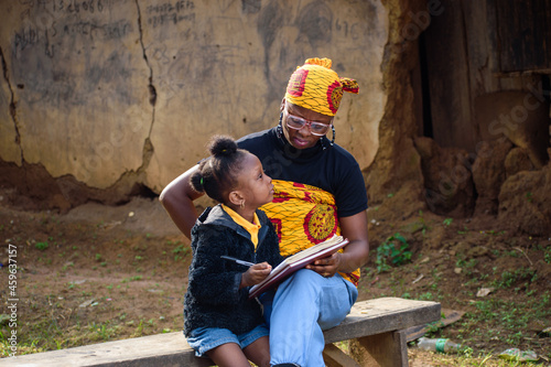 An African mother wearing glasses with a baby on her back, sits outside a village mud house, helping a girl wearing nose mask with studies for excellence in her school, career and education