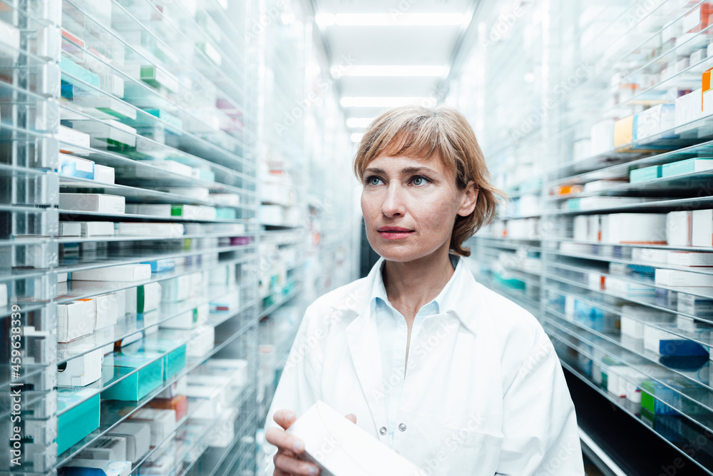 Female pharmacist with medicine looking away while standing by rack at medical store Female pharmacist with medicine looking away while standing by rack at medical store