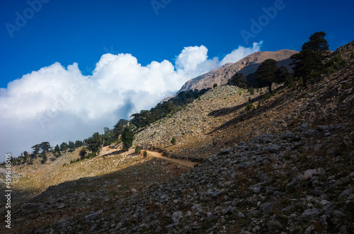 Mountainous landscape, mountainside with stones, sparse vegetation, dry grass, trees in distance and country road. Large fluffy clouds swirling on horizon, Nature of southern Turkey in Antalya region photo