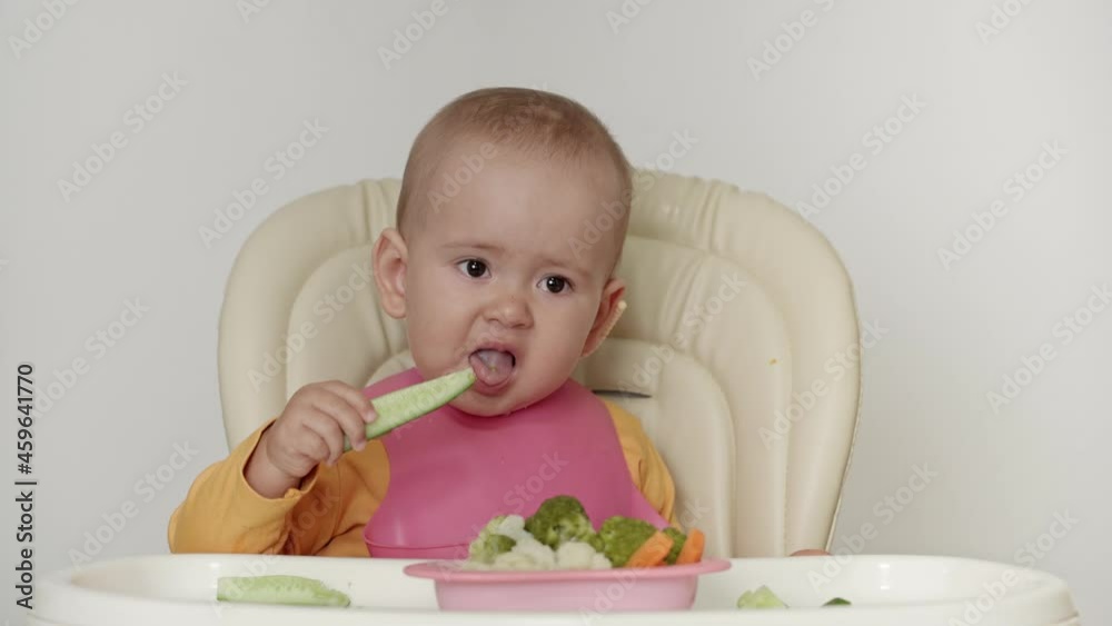 A one-year-old baby is sitting on a high chair. The child tastes a fresh cucumber and refuses to eat it. A one-year-old baby is sitting on a high chair. The child tastes a fresh cucumber and refuses to eat it.