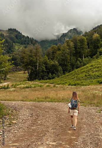 Rear view of young blonde woman with backpack walking uphill along trail on background of mountain covered with forest, active hiking eco tourism
