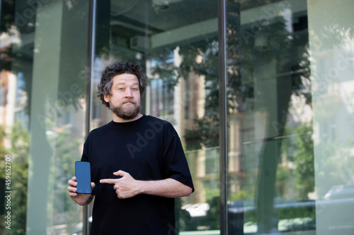 Caucasian man in front of glass building pointing at cell phone screen