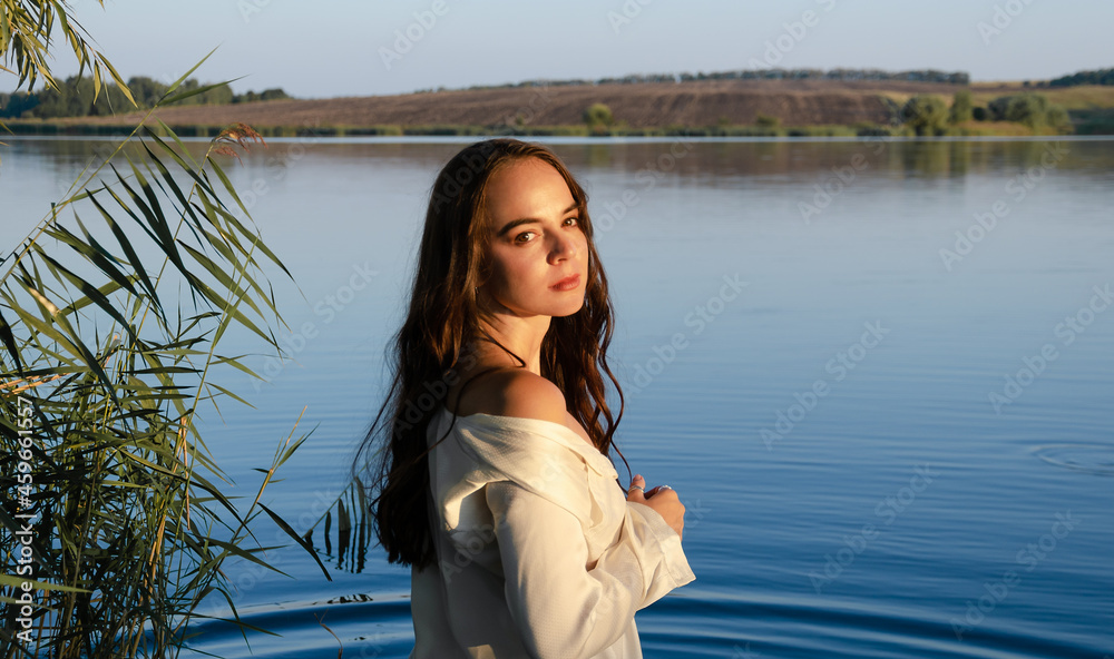Young woman in white shirt stands on the lake reeds shore during sunset, with landscape behind her, looking at camera.