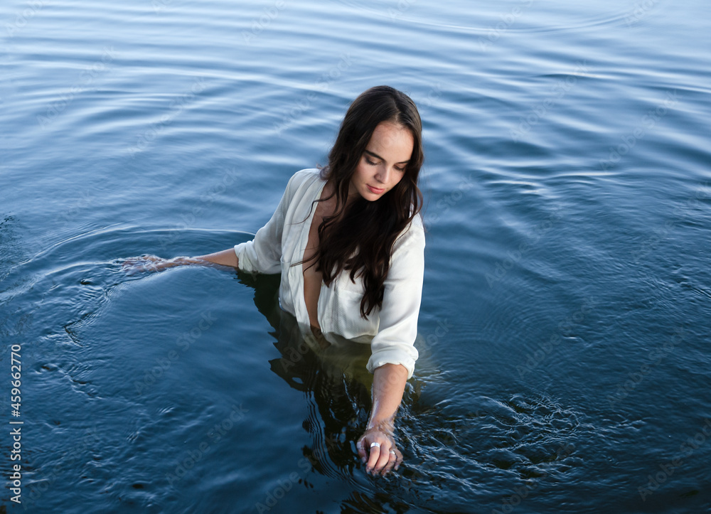 Fototapeta premium Young thoughtful brunette woman in white shirt is standing in the lake and moving hands through water.