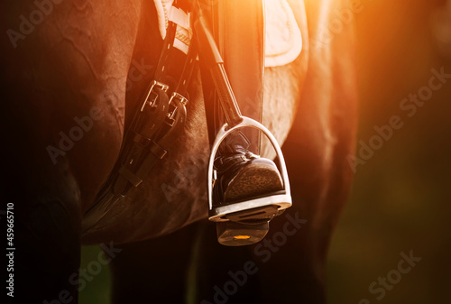 The rider's foot, sitting in the saddle on a horse, is dressed in a black boot and rests on a metal stirrup illuminated by the sun. Equestrian sports. Horse ammunition.
