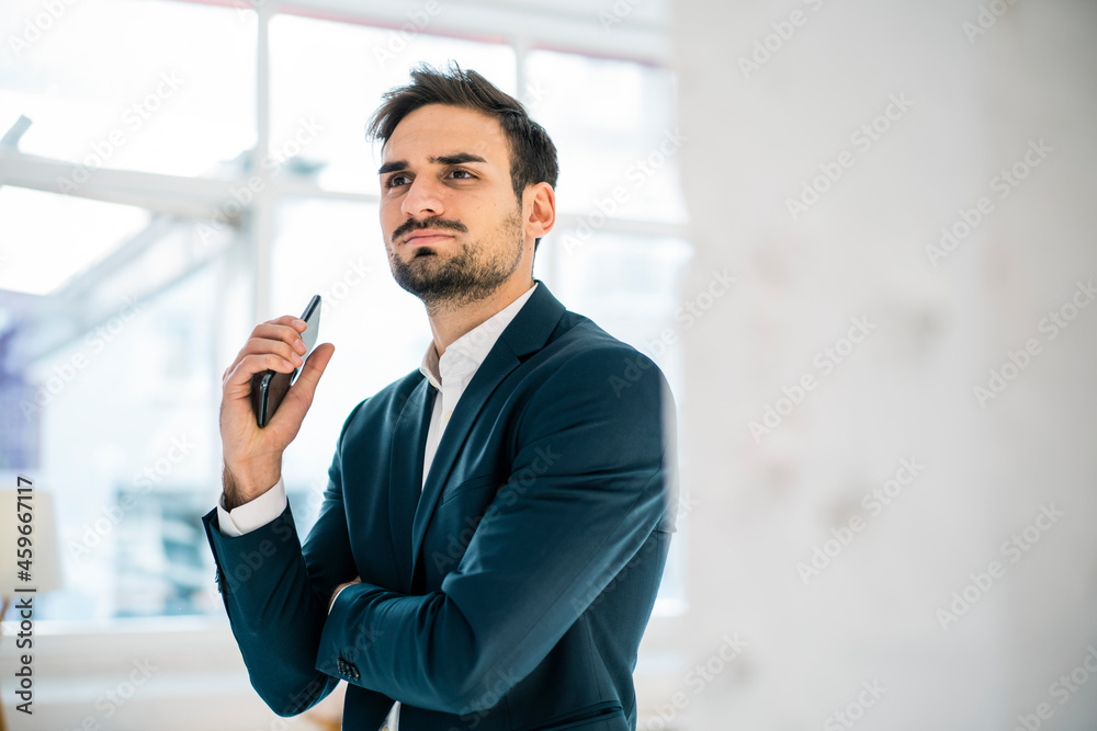 Thoughtful young businessman looking away while holding smart phone in office