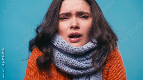 ill woman in knitted scarf sneezing while looking at camera isolated on blue