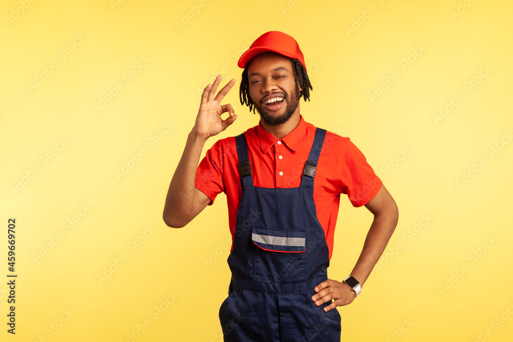 Portrait of sappy handyman with beard wearing overalls showing ok sign, being satisfied of done work in time, recommend service industry. Indoor studio shot isolated on yellow background.