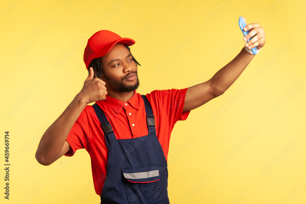 Handsome worker with beard wearing uniform having video call with client, showing thumb up, talking about perfect job, execute order. Indoor studio shot isolated on yellow background.