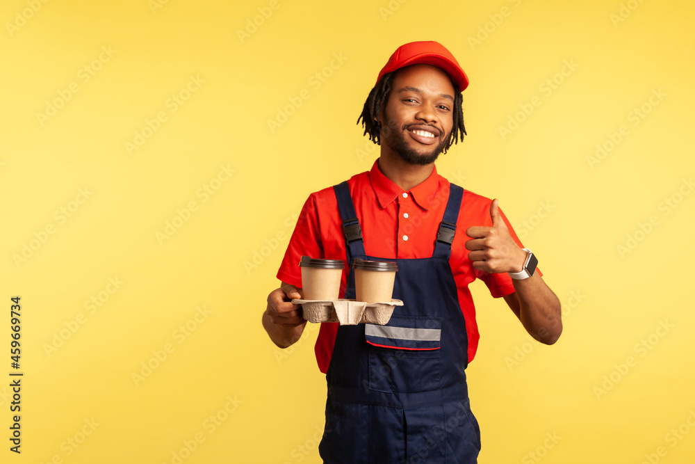 Delivery service. Handsome courier in blue overalls holding coffee, offering drinks in disposable cups and showing thumbs up, like gesture. Indoor studio shot isolated on yellow background.
