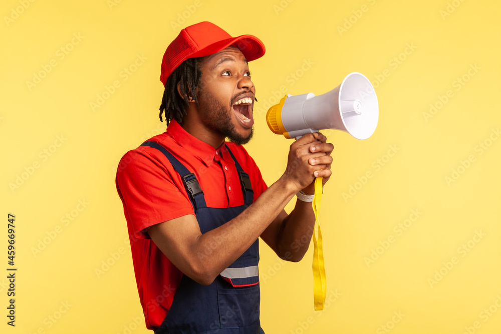 Portrait of bearded handyman wearing blue overalls and red cap screaming announcing about discounts of service industry or protesting. Indoor studio shot isolated on yellow background. Portrait of bearded handyman wearing blue overalls and red cap screaming announcing about discounts of service industry or protesting. Indoor studio shot isolated on yellow background.