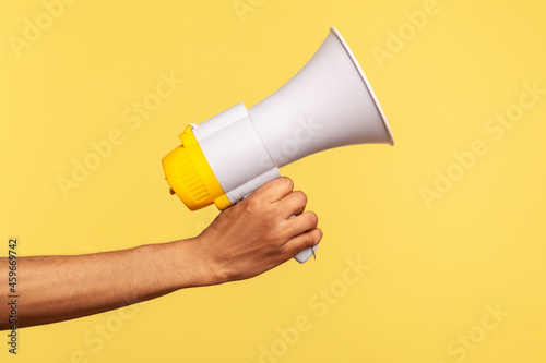 Profile side view closeup of human hand holding megaphone, announcing of advertisement. Indoor studio shot isolated on yellow background.