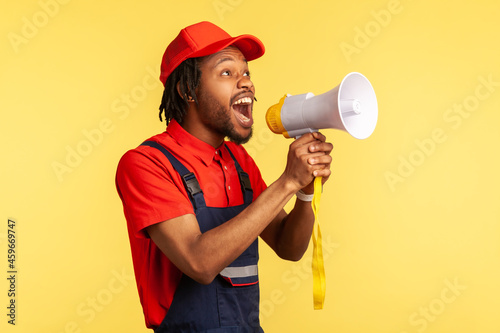 Portrait of bearded handyman wearing blue overalls and red cap screaming announcing about discounts of service industry or protesting. Indoor studio shot isolated on yellow background.