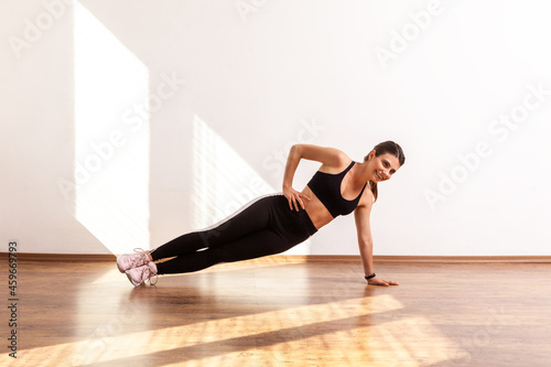 Young adult slim woman in side plank pose with one hand on hip, doing sport exercises at home, wearing black sports top and tights. Full length studio shot illuminated by sunlight from window. © khosrork