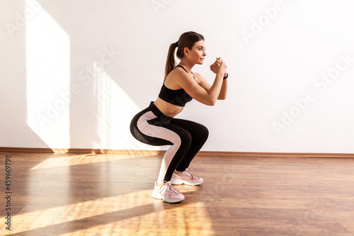 Full length portrait of woman doing split or lunge squat exercise at home or fitness gym, wearing black sports top and tights. Indoor studio shot illuminated by sunlight from window.
