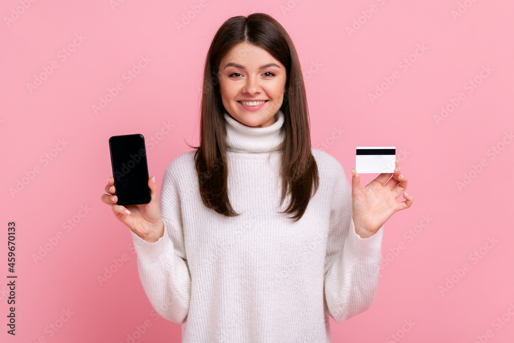 Smiling optimistic female holding credit card and smart phone with empty display for advertisement, wearing white casual style sweater. Indoor studio shot isolated on pink background.