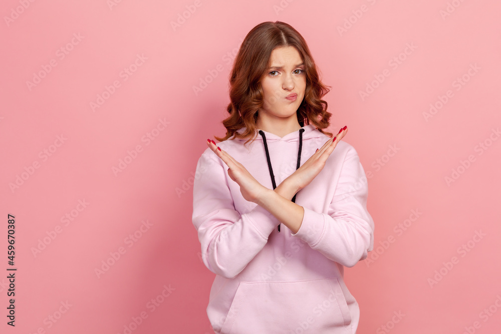 Portrait of serious teen girl with brunette hair in hoodie making x sign with crossed hands, gesturing stop, warning to ban, body language. Indoor studio shot isolated on pink background Portrait of serious teen girl with brunette hair in hoodie making x sign with crossed hands, gesturing stop, warning to ban, body language. Indoor studio shot isolated on pink background