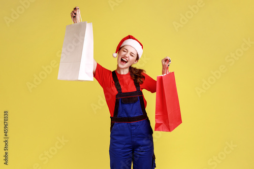 Extremely excited delivery woman holds shopping bags in raised hands, dancing happily, free shipping, wearing blue overalls and santa claus hat. Indoor studio shot isolated on yellow background.
