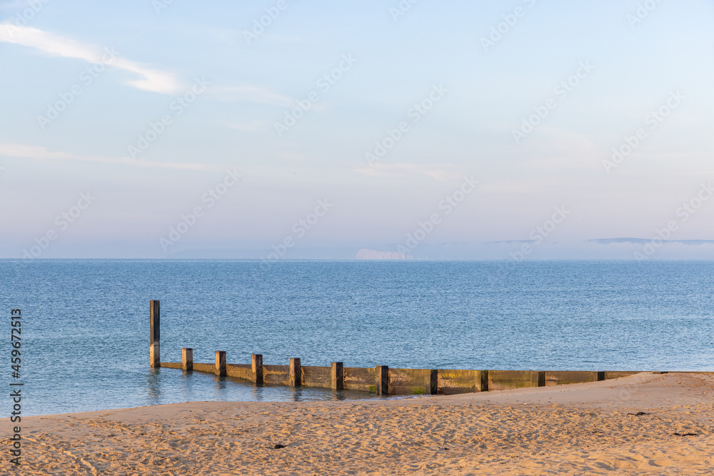 A scenic view of a beautiful blue sea with wooden groyne and some foggy hill in the background under a beautiful blue sky and some white clouds