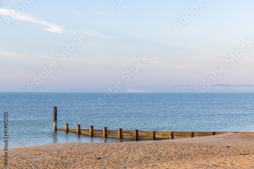 A scenic view of a beautiful blue sea with wooden groyne and some foggy hill in the background under a beautiful blue sky and some white clouds © Dolwolfian