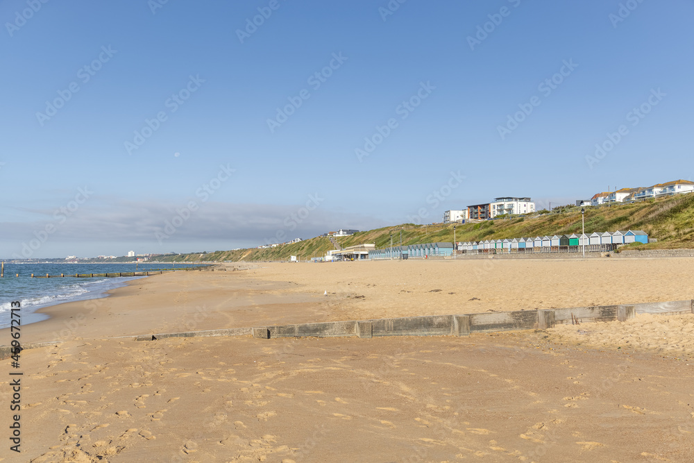 A scenic view of a sandy beach with colorful beach huts, wooden groyne (breakwater) and grassy cliff under a majestic blue sky and some white clouds