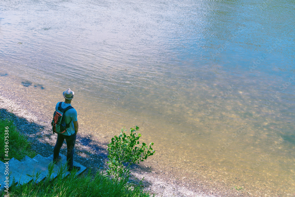 A man in a hat and with a backpack on the nature by the lake looks towards the river