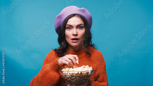 impressed woman holding bowl of popcorn while watching interesting film isolated on blue