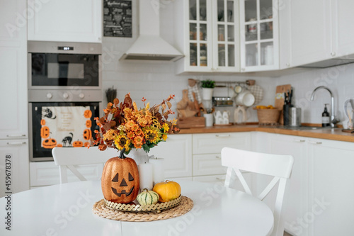 A vase of flowers,a jack pumpkin and candles on a tray. In the background - the interior of a white kitchen in Scandi style. The concept of home and comfort. Autumn decor for the Halloween holiday.