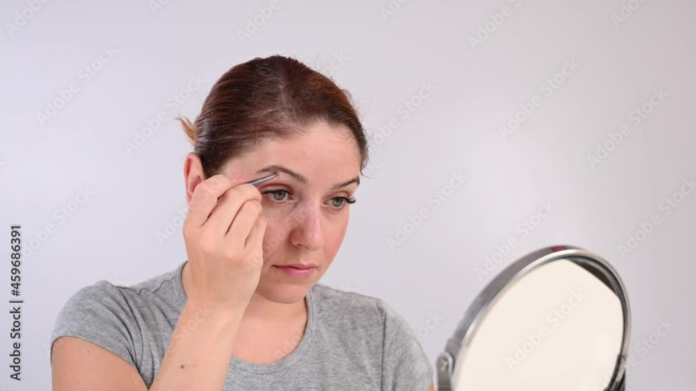 Caucasian woman looks in the mirror and does the eyebrow correction herself with tweezers Caucasian woman looks in the mirror and does the eyebrow correction herself with tweezers