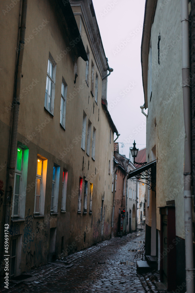 an atmospheric narrow street in the old city of Europe. Old houses, small windows with colored light. A narrow strip of cloudy sky is visible. The houses are three-storied