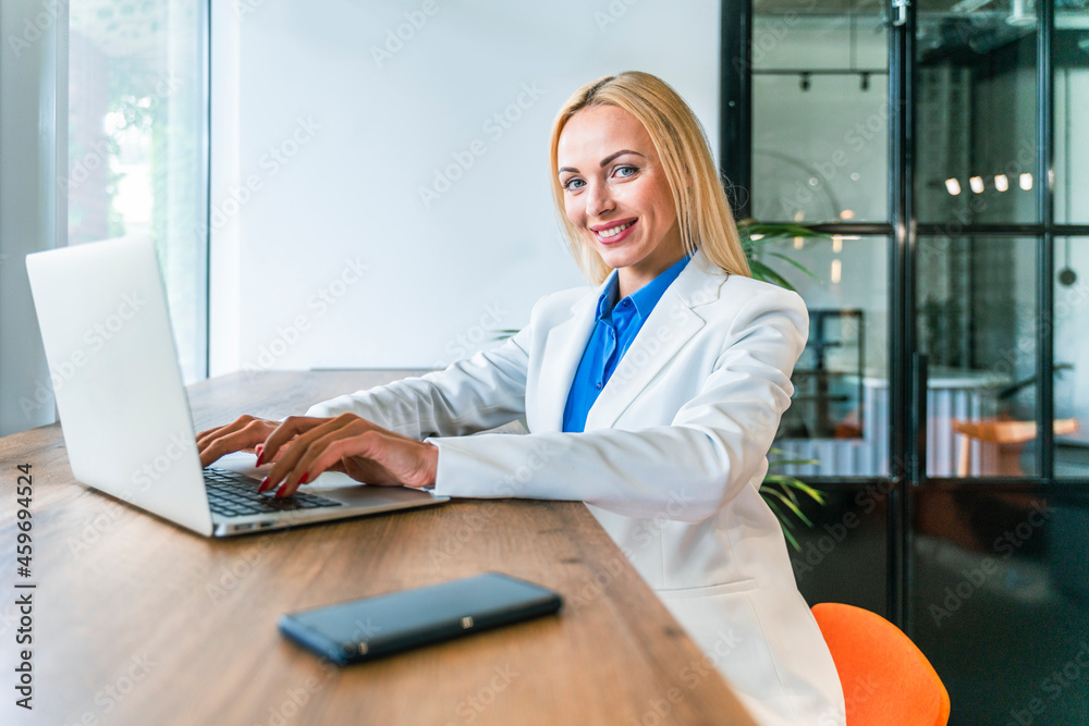 Smiling blond businesswoman with laptop sitting at desk in office Smiling blond businesswoman with laptop sitting at desk in office
