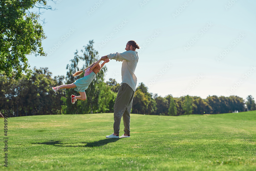 Full length shot of young father and daughter having fun, spinning together on grass field in summer park